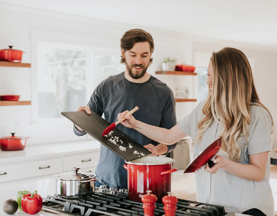 Two people cooking together in a bright home kitchen, one holding a cutting board of chopped onion over a red pot on the stove.
