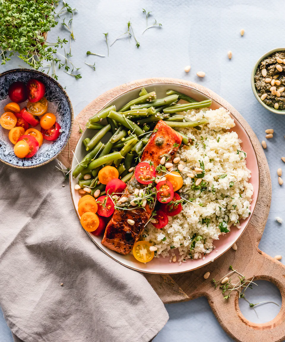 Overhead view of a dinner bowl with glazed salmon, green beans, herbed rice and halved cherry tomatoes on a wooden board, beside a linen napkin.