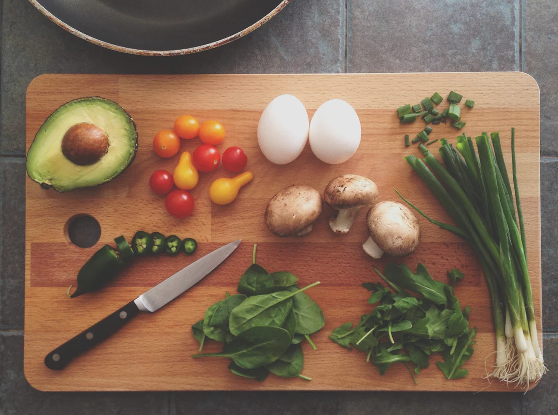 A wooden cutting board laid out with prepped ingredients: half an avocado, cherry tomatoes, eggs, mushrooms, spinach, sliced chili and green onions.