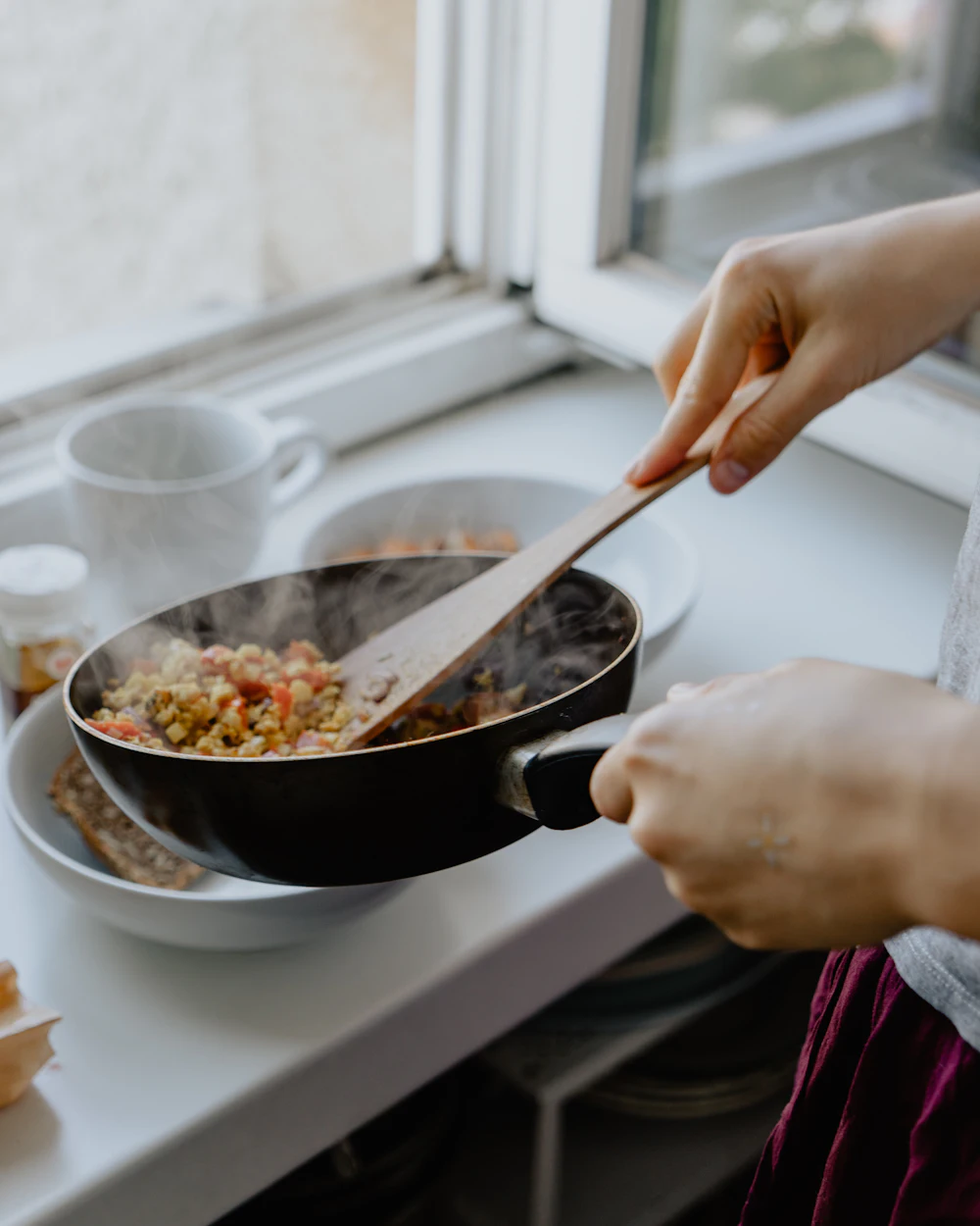Hands stirring a steaming skillet of vegetables with a wooden spoon at a sunny kitchen window.
